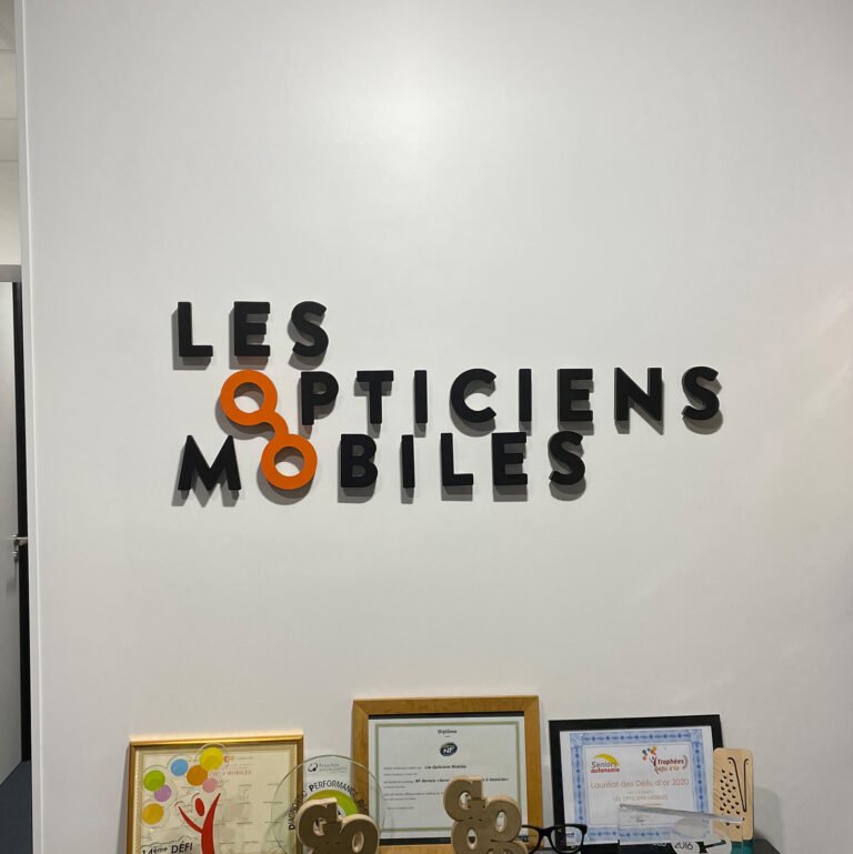 Logo Les Opticiens Mobiles en relief appliqué sur un mur intérieur blanc.